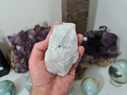 A hand holding a natural Rainbow Moonstone crystal piece that is approximately 10 centimetres by 6 centimetres in size