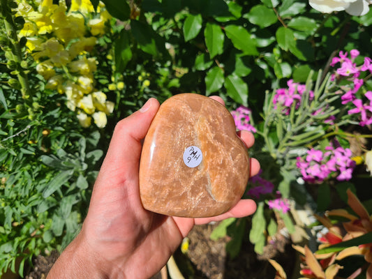 A hand holding a Peach Moonstone crystal heart that is approximately 9.5 centimetres by 9 centimetres in size